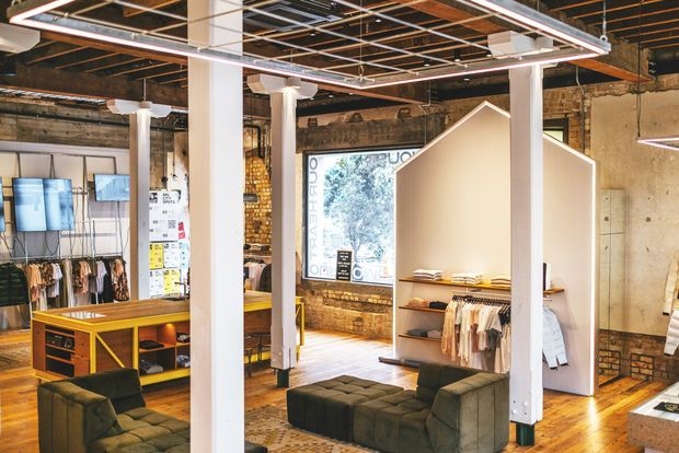 Industrial-style retail showroom or fashion store interior with exposed brick and concrete walls, polished timber flooring, white structural columns, suspended linear lighting, clothing racks, display shelving, counters, and dark upholstered seating. The open plan includes a large shopfront window and a prominent freestanding white display partition.