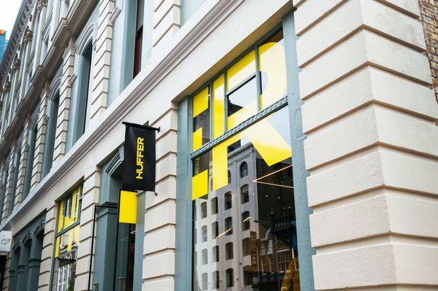 Street-level view of a heritage-style commercial building facade with ornate classical detailing, pilasters, arched windows, and horizontal bands. Large shopfront windows reveal a bold yellow-and-black retail interior and reflect surrounding urban buildings. A vertical black Huffer sign is mounted outside.