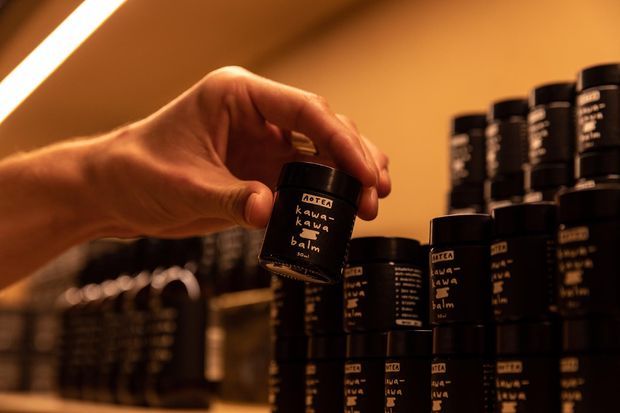 Warmly lit close-up of a hand selecting a small black-labeled product jar from a neatly arranged display of similar jars on retail shelving. The image has an intimate, moody amber atmosphere and appears to show a product display or commercial retail interior.