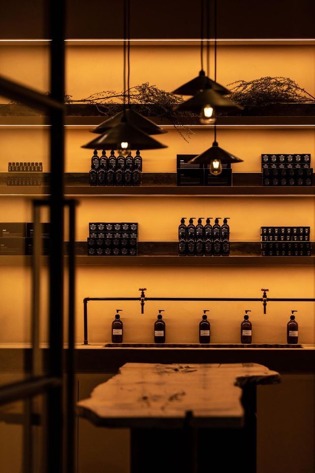 A warmly lit retail or hospitality-style interior with dark wall-mounted shelving displaying rows of bottles and small packaged products. Black pendant lights hang in front of the shelves, while a rustic live-edge timber counter occupies the foreground. The space has a dramatic amber and charcoal palette with a refined, atmospheric presentation.