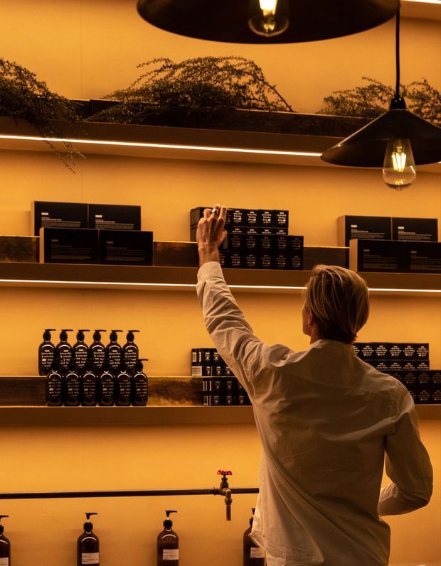 Warm, amber-lit retail interior with dark floating shelves displaying rows of black bottles and boxed products. A person in a light shirt reaches toward the display, while oversized black pendant lights and a large reflective surface add visual depth. The space has a refined, moody boutique atmosphere with integrated shelf lighting and dark stone-like shelf surfaces.