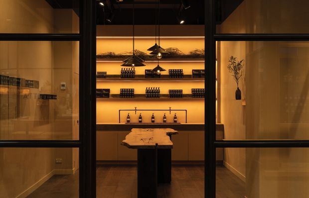 A moody, refined interior view through black-framed glass partitions into a display bar or tasting room. Warm amber backlighting illuminates open shelves holding bottles and glassware, with a dark stone or timber counter island in the centre. The room has dark flooring, black metal framing, recessed ceiling lighting, and a sculptural pendant light.
