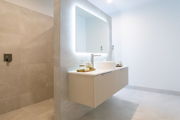 Bright contemporary bathroom featuring a walk-in tiled shower, large backlit wall mirror, floating fluted vanity with a pale benchtop, and light stone-look floor and wall tiles. The palette is soft beige, white, and light grey with clean minimalist detailing.