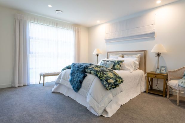 Bright, spacious bedroom with a large upholstered bed dressed in white bedding and layered with a blue floral throw and textured blue blanket. The room has pale walls, grey carpet, sheer floor-to-ceiling curtains, bedside tables with lamps, a cushioned bench, and a decorative wall treatment behind the bed. The overall atmosphere is refined, calm, and inviting.