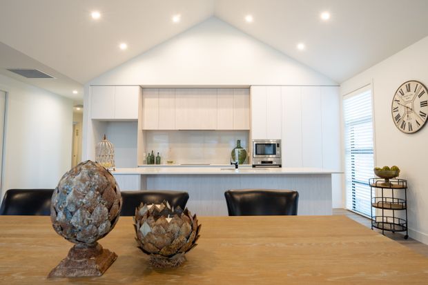 Bright open-plan kitchen and dining area viewed from a timber dining table. The kitchen features a long white island with integrated seating, pale timber cabinetry, a light tiled splashback, stainless-steel appliances, and a dramatic vaulted white ceiling with recessed lighting. A large wall clock, window shutters, and decorative objects add character.