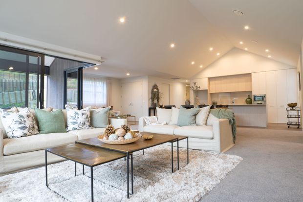 Bright, open-plan contemporary living room flowing into a modern kitchen. The space features large black-framed glazed doors, a vaulted ceiling with recessed lighting, pale upholstered sofas, timber-and-metal nesting coffee tables, a textured light rug, and a light-toned kitchen with a central island.