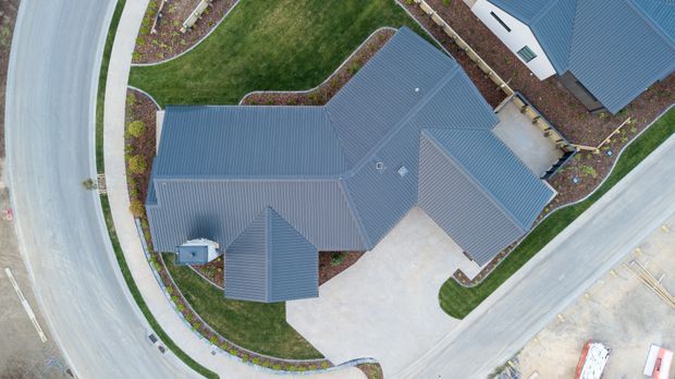 Aerial top-down view of a large residential property showing several interconnected pitched roof forms with dark blue-grey metal roofing, curved concrete driveways and footpaths, landscaped garden beds, and surrounding roads. The image primarily captures the building roofscape and site layout.