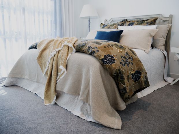 A softly lit bedroom featuring a neatly made bed with layered cream and beige bedding, a patterned floral-botanical throw, and a dark blue accent cushion. The bed has an ornate light-painted cane or wicker-style headboard, with sheer curtains, a floor lamp, and grey carpet contributing to a calm, traditional coastal-inspired interior.