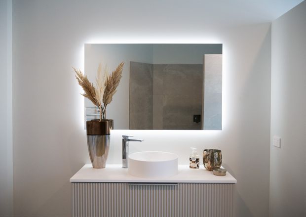 A refined contemporary bathroom vanity featuring a white vessel basin, brushed metal tapware, a fluted built-in vanity cabinet, and a large backlit rectangular mirror. Neutral grey and white finishes create a clean, softly illuminated interior.