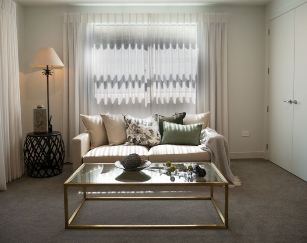 A compact, softly lit living room with a cream upholstered sofa positioned in front of sheer patterned curtains. The room includes a glass-topped rectangular coffee table with a metallic frame, a decorative side table with a table lamp, neutral carpet, pale walls, and a closed white interior door. The styling is refined and neutral with subtle green and patterned cushion accents.