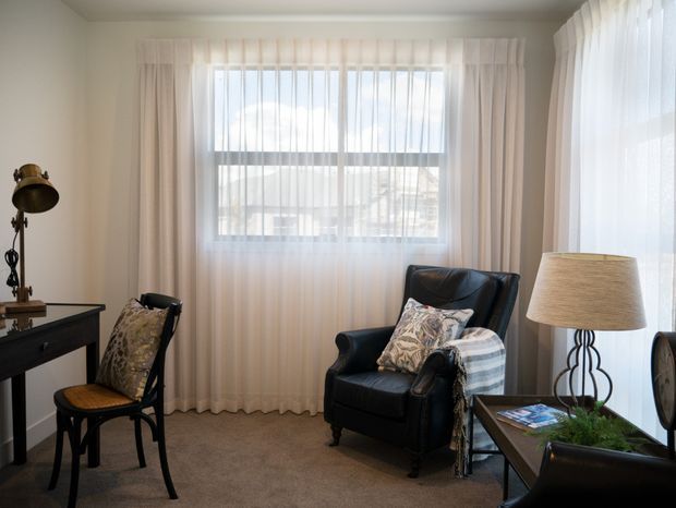 A softly lit traditional sitting room with sheer floor-to-ceiling curtains, a large window, dark leather armchairs, a small side table, a writing desk, and a vintage-style floor lamp. Neutral beige carpeting and pale walls create a calm, comfortable atmosphere.