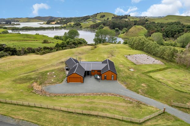 Aerial view of a dark-roofed rural property with orange-brown timber-clad outbuildings, gravel drive, fenced grassy grounds, and a landscaped pond or lake setting amid rolling green hills.
