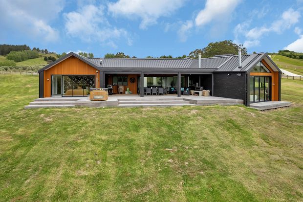 A low-slung contemporary rural house set in a broad grassy landscape. The home has a long linear plan with gabled timber-clad wings, dark brickwork, extensive floor-to-ceiling glazing, a grey metal roof, and a covered outdoor entertaining terrace with seating and a built-in fireplace or barbecue area.