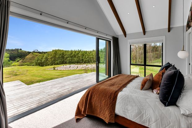 Bright contemporary bedroom with a low-profile bed dressed in white bedding, rust and dark accent cushions, and a textured rust throw. Full-height sliding glass doors open onto a timber deck and landscaped grounds, while a large black-framed window provides additional garden views. The room has a white pitched ceiling with exposed dark timber beams, grey curtains, pale carpet, and a spherical pendant light.