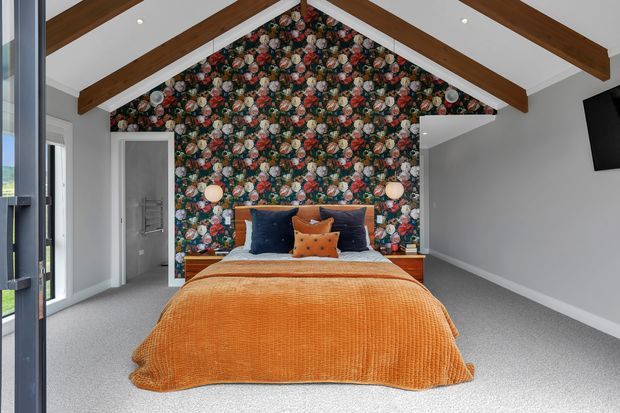 A bright bedroom with a vaulted ceiling and exposed dark timber beams. The bed is centred against a dramatic floral feature wall, with an orange textured bedspread, navy and rust cushions, and grey carpet. White walls and recessed ceiling lights create a clean contemporary backdrop, while a wall-mounted television and an open doorway are visible to the right and left.