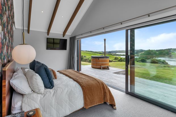 A contemporary bedroom with a low-profile bed, white bedding, layered neutral and navy cushions, and a textured brown throw. The room has grey walls, grey carpet, exposed dark timber ceiling beams, a large abstract wall artwork, pendant lighting, and a wall-mounted television. Full-height sliding glass doors open onto a timber deck with a round outdoor spa or soaking tub, overlooking a green rural landscape and water.