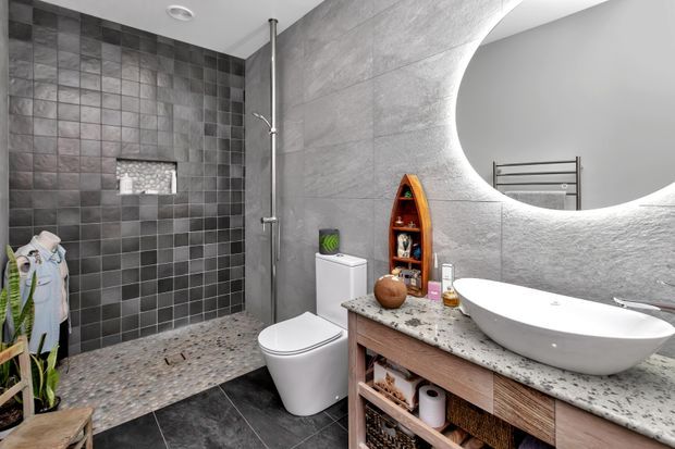 Contemporary bathroom with a walk-in shower clad in dark square tiles, a pebble-style shower floor, wall-mounted shower fittings, a separate toilet, and a freestanding vessel basin on a timber vanity. A large round mirror and curved wall detail create a distinctive feature, while open vanity shelving provides storage.