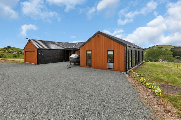 Exterior view of a contemporary single-storey rural home and detached garage with dark metal cladding and warm vertical timber weatherboards. The buildings have simple pitched gable roofs, large gravel forecourt, and landscaped planting, set among rolling green hills under a partly cloudy sky.