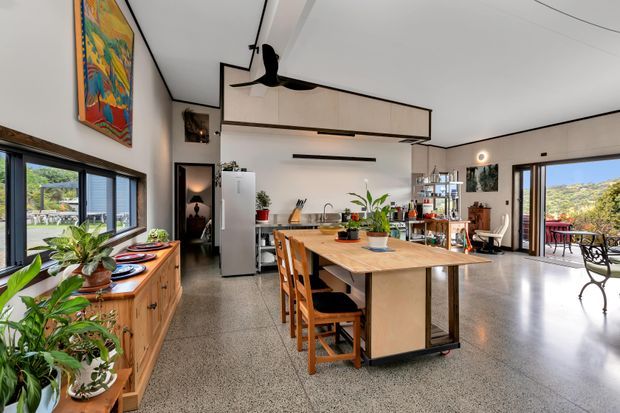 A bright open-plan kitchen and living area with a central timber island, integrated cabinetry, stainless-steel appliances, timber dining stools, extensive windows and sliding doors, and polished concrete or terrazzo-style flooring. The space has a mid-century-influenced feel with indoor plants, colourful artwork, and views to a landscaped outdoor setting.