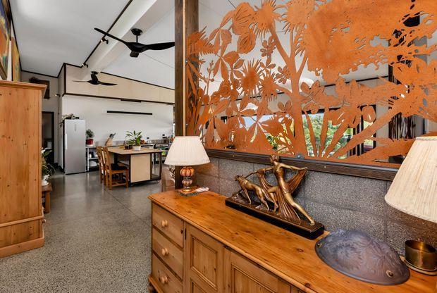 A warmly styled open-plan interior featuring a timber sideboard in the foreground, a large orange botanical metal screen or artwork, decorative lamps and sculptural objects, and a dining and kitchen area beyond. The space has polished grey terrazzo flooring, white ceilings with black trim and ceiling fans, timber cabinetry, and an eclectic blend of natural and vintage-inspired furnishings.