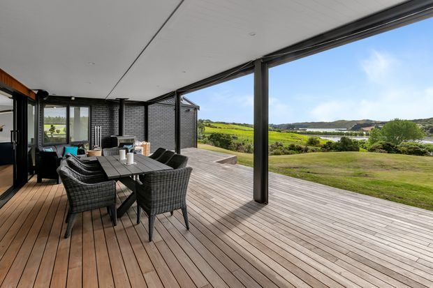 A spacious covered outdoor deck or terrace with timber decking, a dining table and woven chairs, and a large open structural frame overlooking a green rural landscape and water. The contemporary pavilion-like extension has dark-framed glazing, a flat white ceiling, and dark brick walls, creating a sheltered indoor-outdoor entertaining area.