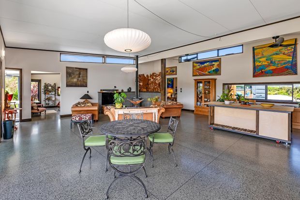 Open-plan living and dining area with polished concrete flooring, white walls, high ceilings, clerestory windows, and a large sculptural pendant light. A round metal dining table with green upholstered chairs sits in the foreground, while leather seating, timber cabinetry, artwork, and a kitchen or bar zone occupy the background.