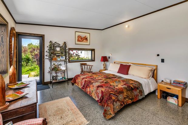 A warmly styled bedroom with a low timber-framed bed, patterned red and orange bedspread, bedside table, floor rug, timber furniture, plants, artwork and shelving. The room has white walls and ceiling with dark timber trim, polished concrete or terrazzo-like flooring, and a glazed door opening to a garden.
