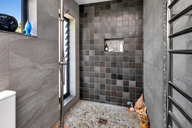 Contemporary walk-in shower with dark grey square wall tiles, a pebble mosaic shower floor, recessed niche, frameless glass screen, and a chrome heated towel rail. A narrow louvred window provides natural light, while small decorative objects sit near the shower floor.