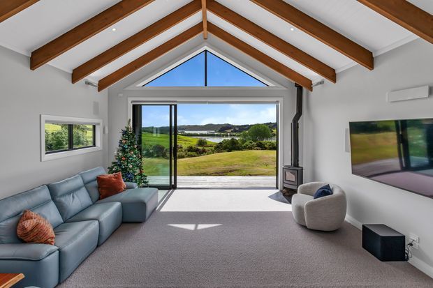 Bright contemporary living room with a vaulted ceiling and exposed timber rafters. The room features a large blue sectional sofa, two rounded lounge chairs, a wall-mounted television, a compact wood-burning stove, and a full-height glazed opening looking across a green rural landscape and water. A Christmas tree adds a seasonal accent.