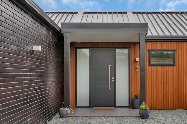 Contemporary residential entrance with a dark metal-clad gabled roof, charcoal brick wall, timber-lined facade, and a sheltered porch framing a modern dark front door with frosted sidelights. Potted plants flank the entry over a gravel forecourt.