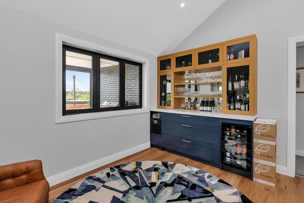 A stylish home bar and beverage storage area in a compact interior room. The built-in joinery combines dark navy lower cabinetry with warm timber-framed glass display cabinets, open shelving stocked with glassware and bottles, a sink or preparation zone, and under-counter wine refrigeration. Black-framed windows bring in daylight and reveal a brick exterior wall beyond. The room has light grey walls, timber flooring, recessed ceiling lighting, and a bold geometric area rug, with part of a brown leather chair visible in the foreground.