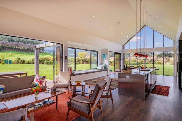 A bright, open-plan contemporary living, dining and kitchen space with expansive floor-to-ceiling glazing overlooking a green rural landscape. The room has a dramatic high pitched ceiling, warm timber flooring, a streamlined kitchen island, mid-century-inspired lounge furniture, and a cluster of red pendant lights above the island.