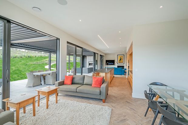 Bright contemporary open-plan living and dining area with floor-to-ceiling glazing opening onto a covered outdoor patio and lawn. The room features a grey sofa with red cushions, timber coffee tables, a glass dining table with dark chairs, warm timber herringbone flooring, white walls and ceiling, and a timber kitchen with island cabinetry in the background.