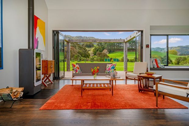 A bright mid-century-inspired living room with dark timber flooring, a large orange-red area rug, and a sculptural black fireplace at left. A low timber coffee table, lounge seating, and colourful cushions face broad openings to a covered outdoor terrace and expansive green rural landscape. Large glazed doors and windows bring the landscape into the room, while a bold abstract artwork adds colour.