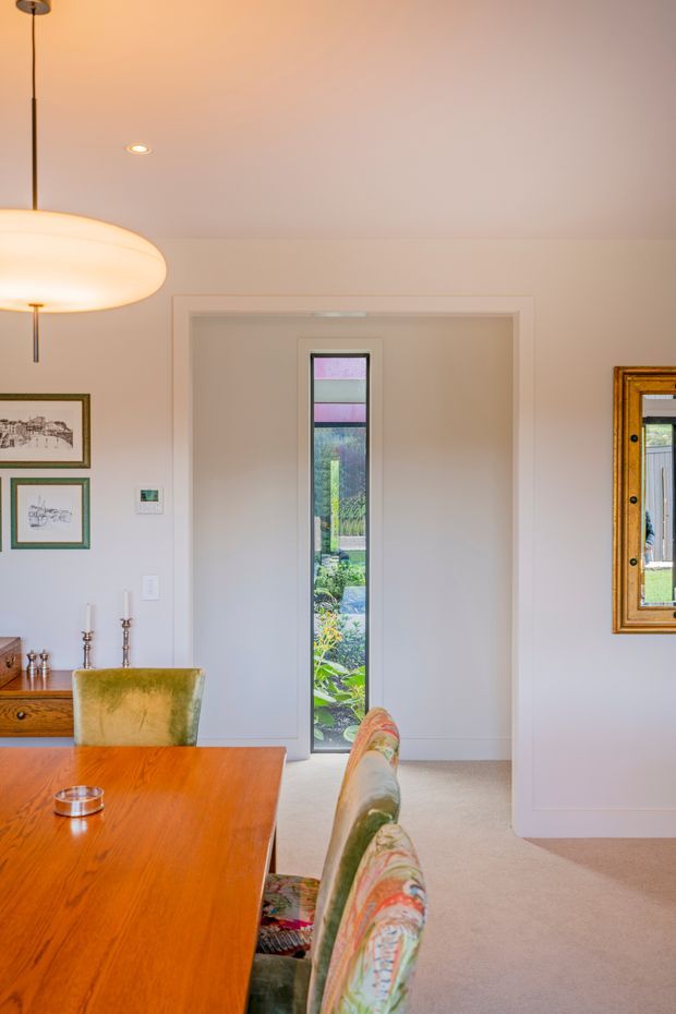 A bright, refined dining room with a long warm timber dining table, upholstered dining chairs, white walls and carpet, framed artwork, a large ornate mirror, and a narrow glazed door opening to a garden. A contemporary pendant light and recessed ceiling lighting illuminate the space.