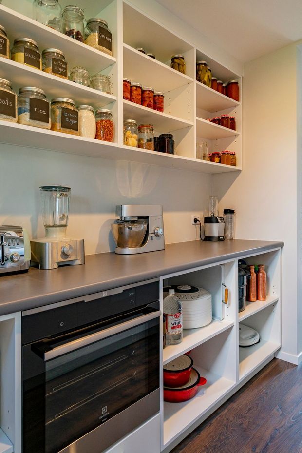 A compact, well-organised pantry or kitchen storage area with open white shelving filled with labelled glass jars, preserves and dry goods. A grey countertop runs above built-in black appliances and open lower shelving containing cookware, bottles and small appliances. The space has white walls and joinery, warm timber flooring, and practical food-preparation equipment including a mixer, blender and toaster.