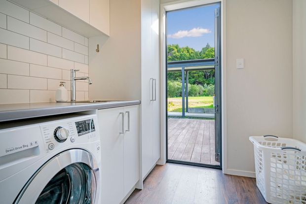 Bright, compact laundry room with white built-in cabinetry, a front-loading washing machine, white subway-tile splashback, stainless-steel tapware, and warm timber flooring. A full-height glazed door opens directly onto a timber deck and garden beyond. A white laundry basket sits in the foreground.