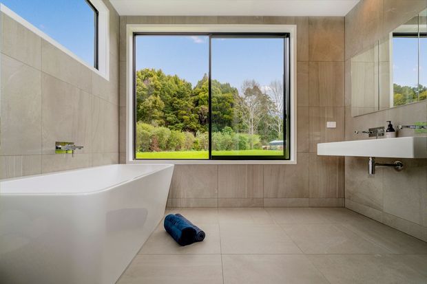 Contemporary bathroom with a freestanding white bathtub, wall-mounted white basin, black tapware, and large-format beige-grey tiles. A large window overlooks a leafy garden, creating a bright and clean atmosphere.