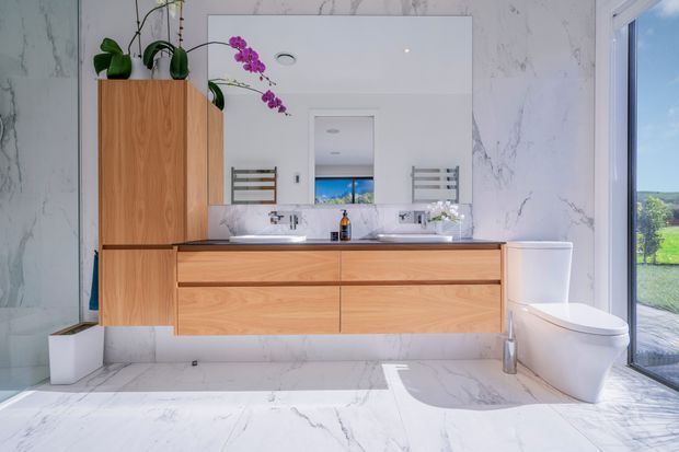 Bright contemporary bathroom with a floating timber vanity, double basins, large frameless mirror, white freestanding bathtub, wall-hung toilet, and dramatic white marble-look wall and floor finishes. A large glazed opening provides views to greenery outside, while orchid plants add a small decorative accent.