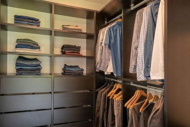 A built-in walk-in wardrobe with dark timber-toned cabinetry, open shelving, drawers, and hanging rails. Folded clothing is arranged on the shelves, while shirts, jackets, and trousers hang on wooden and metal hangers. Warm integrated lighting illuminates the compact, highly organized storage space.