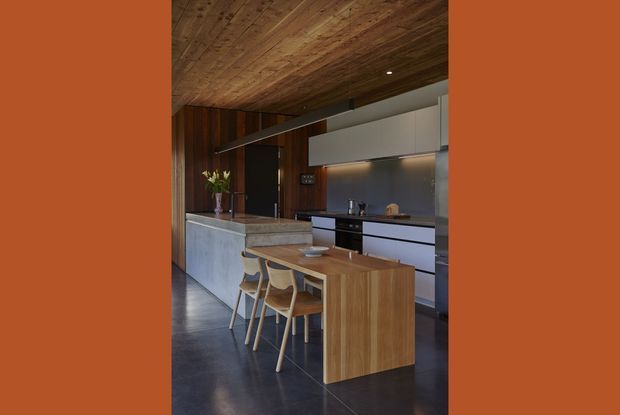 A refined contemporary kitchen with a large stone island and integrated timber dining extension, paired with sculptural timber bar stools. Dark timber wall and ceiling panelling create a warm, enveloping atmosphere, contrasted by pale cabinetry, a stainless-steel backsplash, and dark polished flooring. Minimal linear pendant lighting and concealed under-cabinet illumination complete the streamlined fit-out.