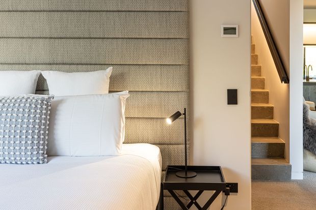 A refined bedroom interior with a large upholstered, horizontally panelled bedhead in a neutral textured fabric, crisp white bedding, and patterned grey-blue accent cushions. A compact black bedside table with a task lamp sits beside the bed. To the right, a carpeted staircase with a dark handrail leads upward, with a glimpse of an adjacent living area beyond. The palette is soft and neutral with tailored, contemporary detailing.