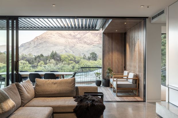 A refined contemporary living room opening onto a covered outdoor terrace, with floor-to-ceiling glazing framing a dramatic mountain and landscaped valley view. The room features a large neutral sectional sofa, dark coffee table, timber slat feature wall and ceiling, timber-framed lounge chairs, polished concrete flooring, and slim dark window and door frames. A pergola-like louvred roof extends over the terrace.