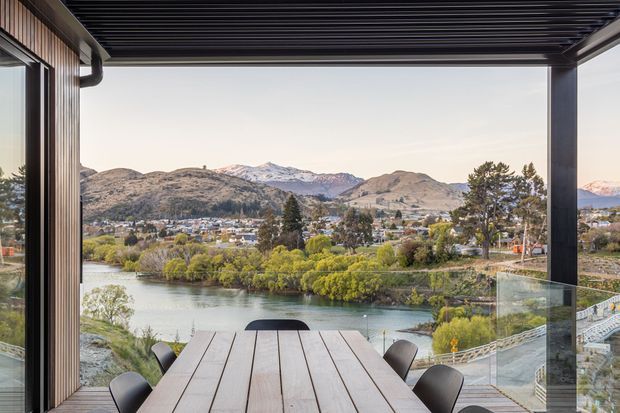 A covered outdoor dining terrace overlooks a calm river, landscaped riverbanks, a town, and surrounding rugged mountains. The space features a long timber dining table, dark chairs, clear glass balustrading, vertical timber wall cladding, and a dark slatted overhead roof structure. The view and open-sided enclosure create a contemporary, scenic entertaining area.