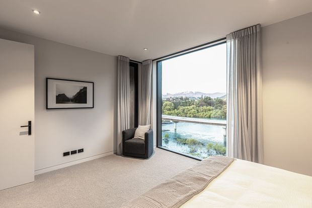 A bright contemporary bedroom with a large floor-to-ceiling window overlooking a landscaped waterfront or river setting and distant mountains. The room has light neutral walls, carpeted flooring, a low-profile bed with a textured beige throw, a dark upholstered armchair, full-height grey curtains, recessed ceiling lights, and a minimalist framed artwork. A partially open door and black hardware are visible on the left.
