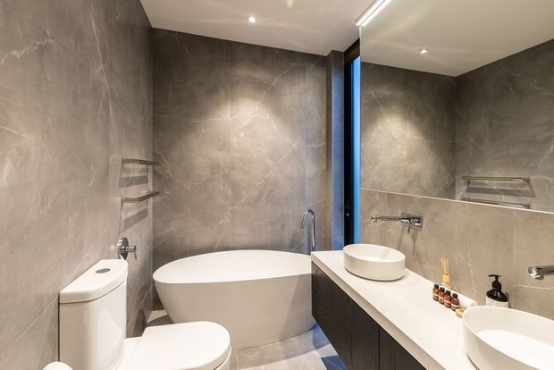 A compact contemporary bathroom with full-height grey stone-look wall tiles, a freestanding white bathtub, wall-mounted fixtures, a white toilet, and a dark timber vanity with white countertop and round vessel basins. A large mirror reflects the vanity and bath area, while recessed ceiling lights provide warm, even illumination.
