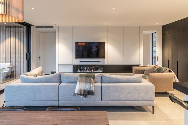 A contemporary open-plan living room viewed from behind a large pale grey sectional sofa. The room features a wall-mounted television set into white full-height cabinetry, a dark built-in media console and cabinetry wall, a warm timber dining table in the foreground, a beige armchair, recessed ceiling lights, and a glazed/slatted opening along the left side. The palette is neutral and refined with timber and dark joinery accents.
