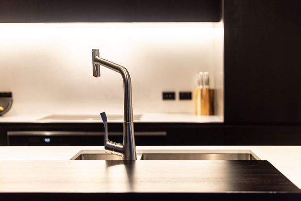 Close-up view of a contemporary kitchen sink and tall brushed metal mixer tap, set into a dark benchtop with a light backsplash and dark cabinetry. Warm under-cabinet lighting creates a clean, refined, high-contrast composition.