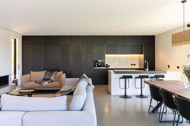 Open-plan contemporary kitchen, dining and living area with a large dark timber-toned built-in wall, integrated kitchen cabinetry and illuminated splashback, white island with black stools, pale upholstered sofas, and a timber dining table. The space is bright and refined with a restrained neutral palette and polished concrete or stone-like flooring.