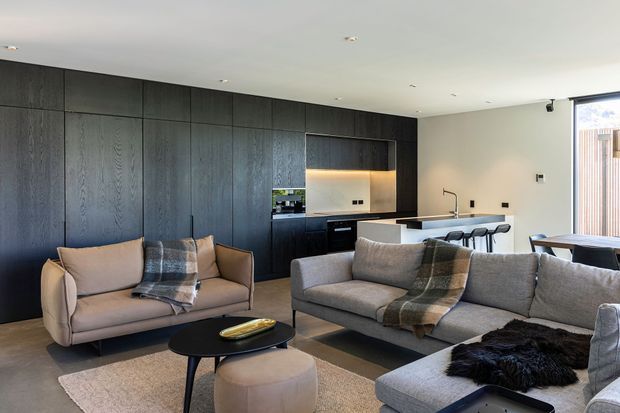 Open-plan contemporary living room with a large grey sectional sofa and separate armchair arranged around a low black coffee table, facing a dark timber-panelled kitchen wall. The kitchen includes integrated black cabinetry, a light stone-look benchtop, warm under-cabinet lighting, and a compact island with bar seating. Pale walls, recessed ceiling lights, neutral textiles, and large glazing create a refined, warm-toned interior.